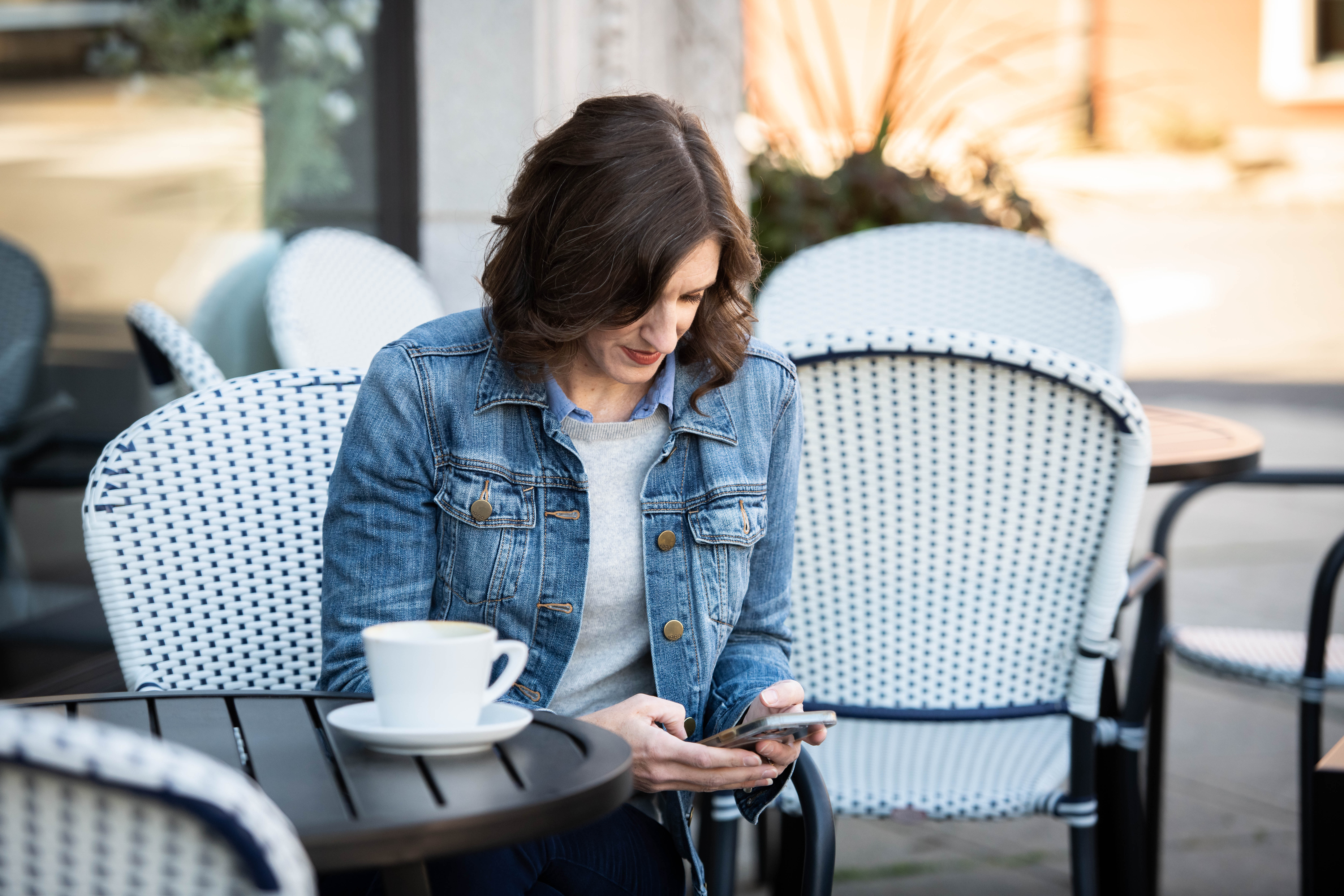 Solopreneur Annie Figenshu working remotely at an outdoor café, checking her phone with a cup of coffee on the table.