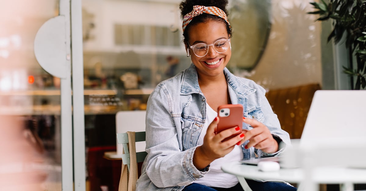 Smiling woman sitting at a café table using her smartphone, with a laptop and coffee nearby