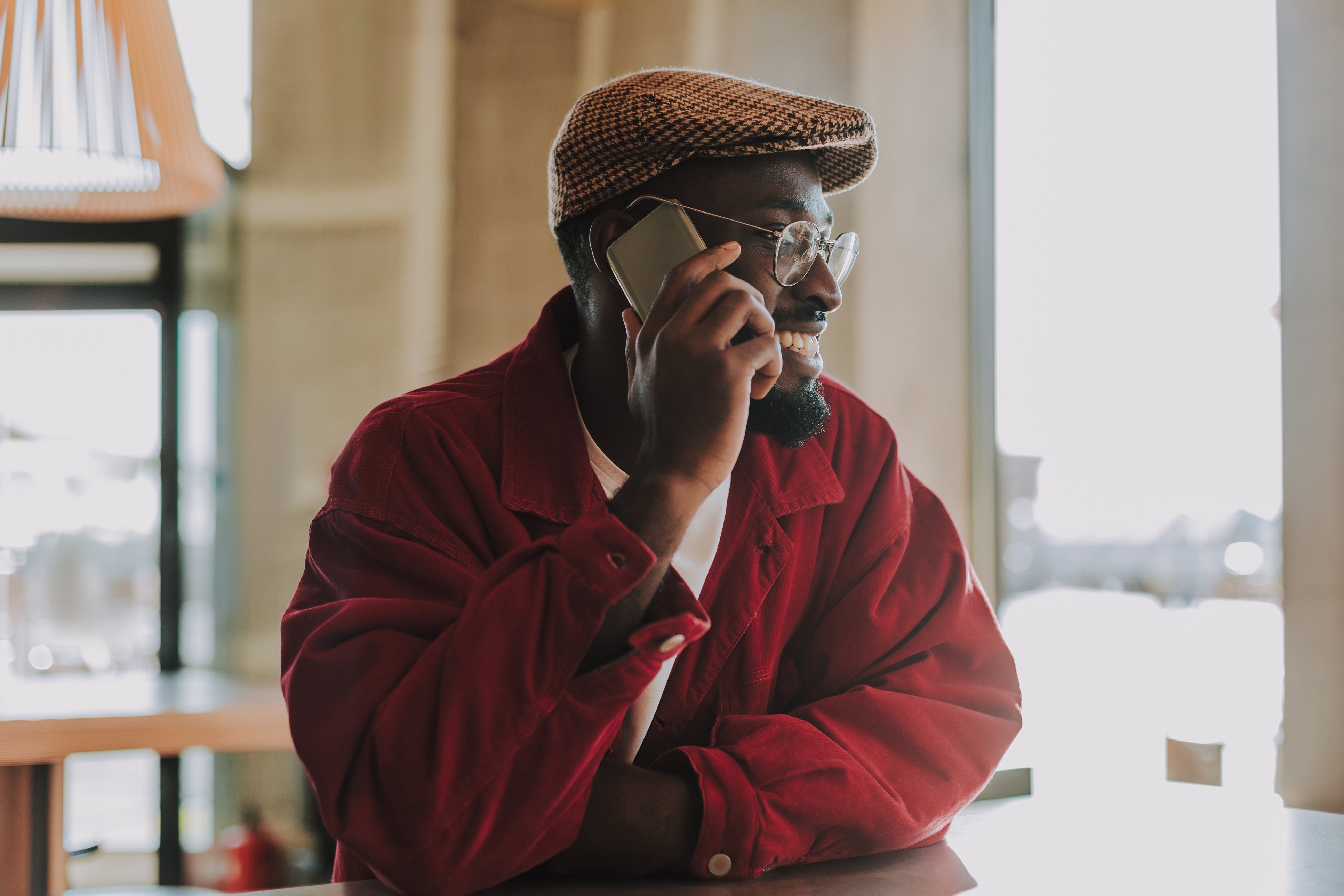Smiling man in a red jacket and houndstooth cap talking on a smartphone in a sunlit café.