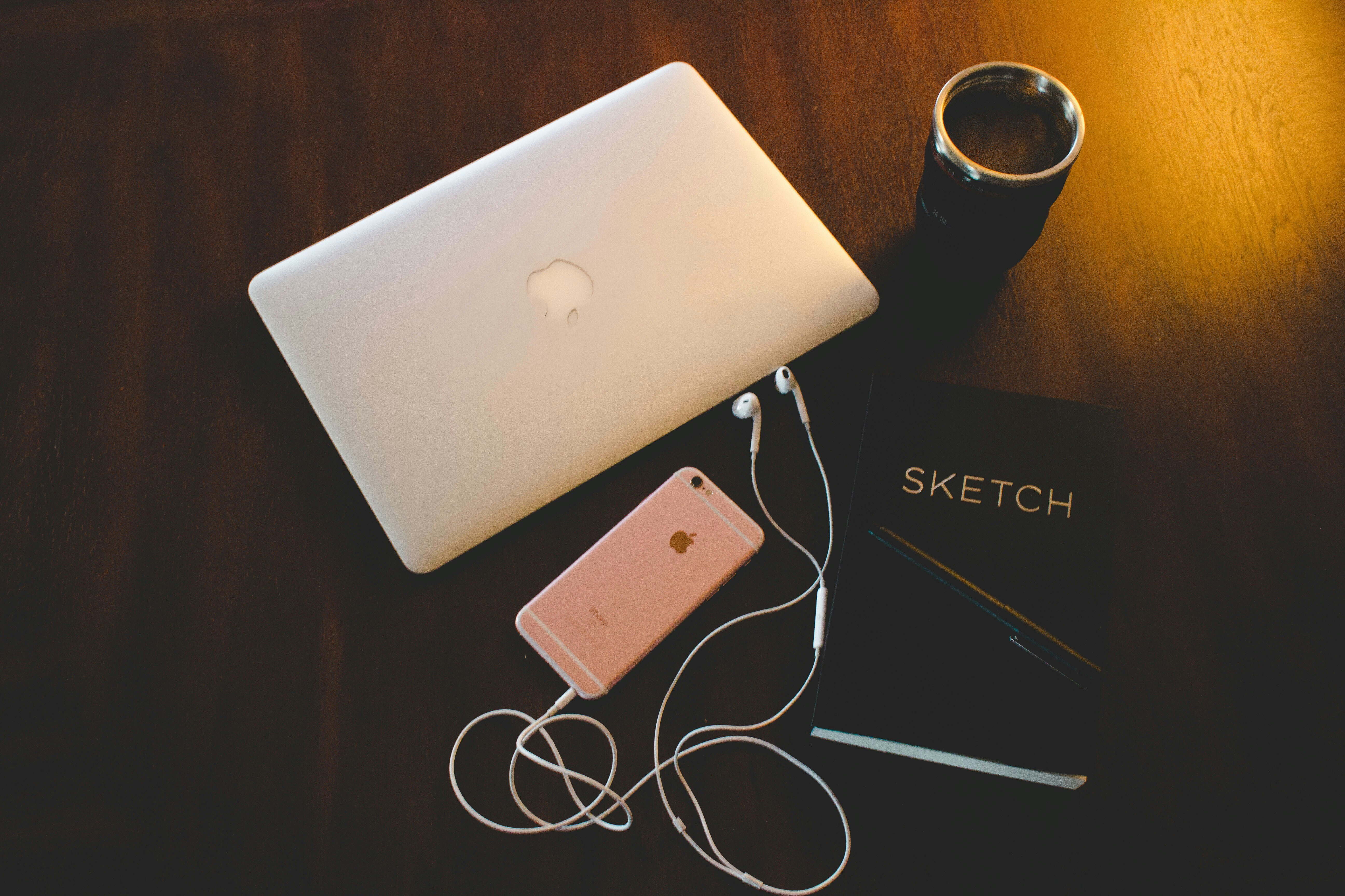 A laptop, phone, and headphones sit on a table with a sketchbook.